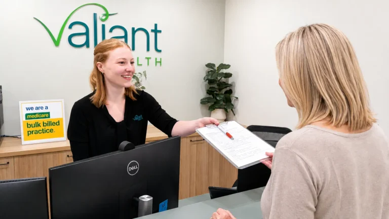 Friendly Valiant Health receptionist handing a clipboard to a patient at a modern Medicare bulk billed medical practice reception in Port Macquarie.